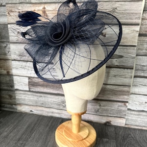 May include: A navy blue fascinator hat with a floral design and netting. The hat is displayed on a mannequin head with a wooden stand. The hat features a circular base and is adorned with feathers.