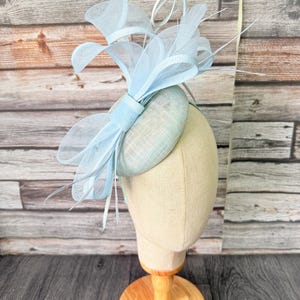 Könnte beinhalten: Ein hellblauer Fascinator, präsentiert auf einem Schaufensterkopf. Das Kopfstück hat eine runde Basis, verziert mit zarten Netzen, Federn und Schleifen. Das Design ist ein weiches, pastellfarbenes Blau, passend für besondere Anlässe.