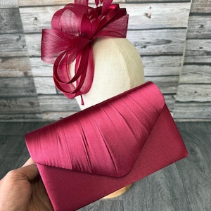 Pode incluir: Uma bolsa clutch de cetim em tom bordô profundo com aba plissada, acompanhada por um fascinator bordô a condizer com laços estruturados e penas delicadas, ambos exibidos num manequim de tom neutro.