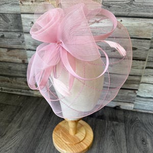 May include: A pale pink fascinator featuring a rose-shaped detail and fine netting. The headpiece is presented on a wooden stand. It incorporates ribbons and feathers, offering a delicate and refined appearance. Ideal for formal events and celebrations.