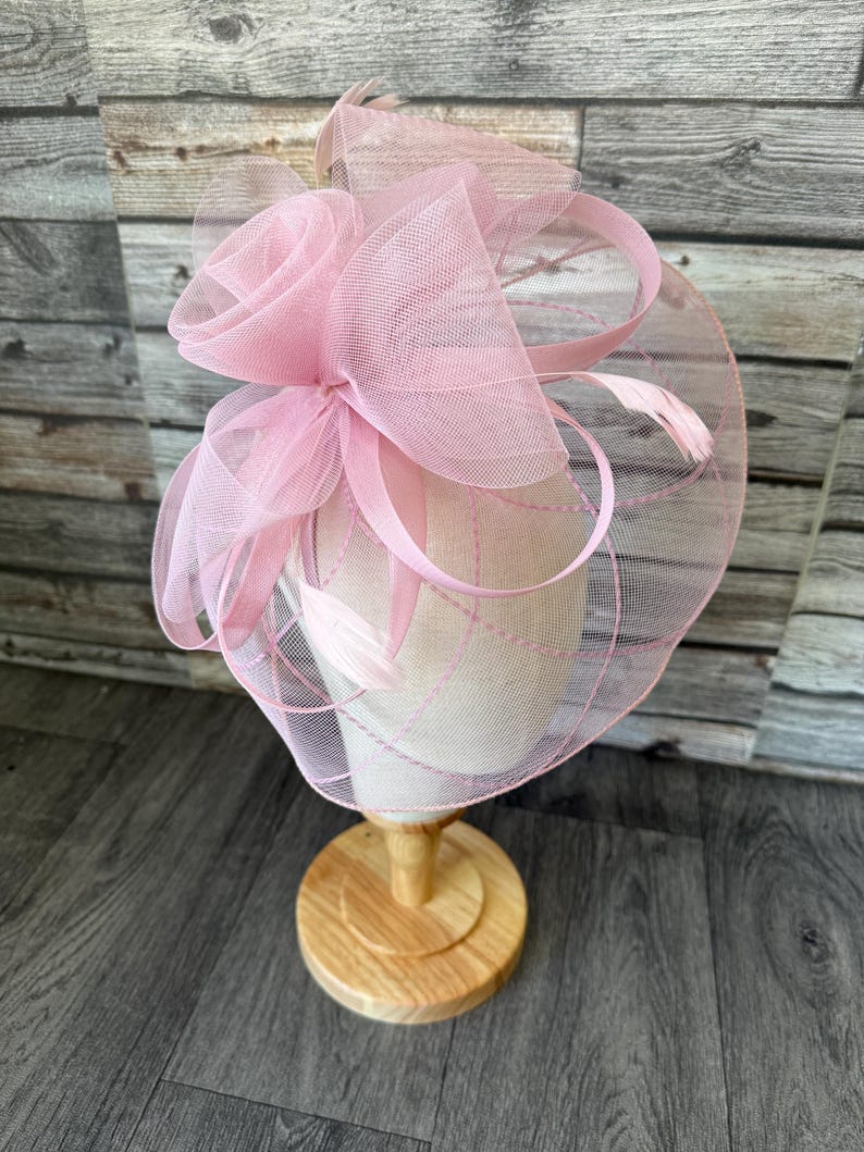 May include: A pale pink fascinator featuring a rose-shaped detail and fine netting. The headpiece is presented on a wooden stand. It incorporates ribbons and feathers, offering a delicate and refined appearance. Ideal for formal events and celebrations.