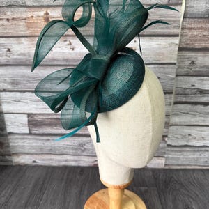 May include: A dark teal fascinator with a structured design, featuring looped accents and feathers. The headpiece is displayed on a mannequin head with a wooden stand, set against a wood-panelled backdrop.