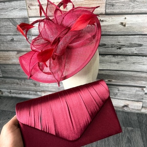 Pode incluir: Um chapéu formal bordô com detalhes de penas e uma clutch a condizer. O chapéu apresenta um design de malha e é montado numa cabeça de manequim. A clutch tem um design plissado.