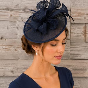 May include: A navy blue fascinator hat with a round base and decorative mesh and feather accents. The hat is worn by a person wearing a navy blue dress. The hat is a formal accessory.