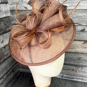 May include: A brown formal hat with a wide brim and decorative accents. The hat features a woven texture and is adorned with feathers and a large bow. The hat is displayed on a mannequin head with a wooden stand.