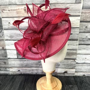 Pode incluir: Um chapéu formal bordô com aba larga e penas decorativas. O chapéu é feito de um material tecido e é exibido em uma cabeça de manequim. O chapéu é de cor vermelho escuro e é adequado para ocasiões especiais.