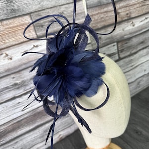 Könnte beinhalten: Ein marineblauer Fascinator mit einem Federbüschel und dekorativen Schlaufen. Der Kopfschmuck wird auf einem Mannequinkopf präsentiert, um sein Design und seine Struktur zu zeigen. Die Federn sind so angeordnet, dass sie voluminös wirken.