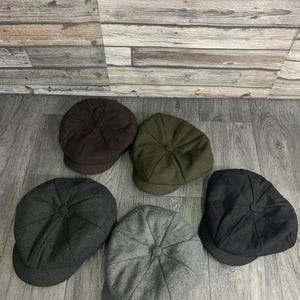 May include: Five newsboy caps in different shades: dark brown, olive green, black, charcoal grey, and light grey. Each cap has a rounded crown with a button detail and a short, curved brim. The caps are displayed on a wooden surface.
