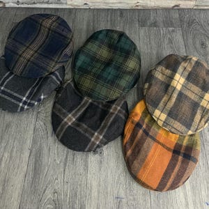 May include: A collection of six plaid newsboy caps in various colors. The caps feature classic patterns in shades of blue, green, brown, and orange. The caps are arranged on a wooden surface.