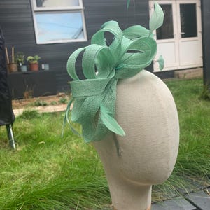 Peut inclure: Un fascinateur vert menthe avec des boucles de tissu et des plumes, présenté sur une tête de mannequin. La coiffe présente une texture tissée et est conçue pour les événements formels. L'arrière-plan comprend une structure en bois et de l'herbe verte.