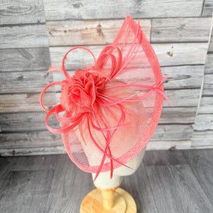Puede incluir: Un fascinador de color coral con una base de malla y detalles decorativos de plumas y flores. El tocado se exhibe sobre una cabeza de maniquí y un soporte de madera, mostrando su diseño y estructura.