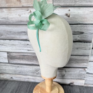 Könnte beinhalten: Ein mintgrüner Fascinator mit Schleife und Federakzenten. Das Kopfstück wird auf einem Schaufensterpuppenkopf mit Holzständer präsentiert. Die Schleife besteht aus einem strukturierten Stoff, die Federn sind weich.