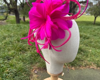 Könnte beinhalten: Ein leuchtend pinkfarbener Fascinator mit einer großen, federartigen Blume und Schleifenakzenten. Der Kopfschmuck wird auf einem Schaufensterpuppenkopf mit Holzständer vor verschwommenem Hintergrund präsentiert. Dieses Accessoire ist für besondere Anlässe geeignet.