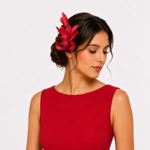 May include: A red hair accessory, possibly a fascinator or clip, is worn on the side of the head. The accessory features a layered design with a feather-like appearance. The woman in the image is wearing a red dress.