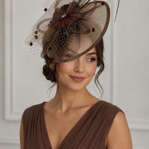 Peut inclure: Un bibi marron foncé composé d'une base circulaire large, d'une fleur en tissu centrale et d'un voile en résille délicat orné de petits accents circulaires sombres. L'accessoire comprend une fine tige incurvée s'étendant vers le haut.