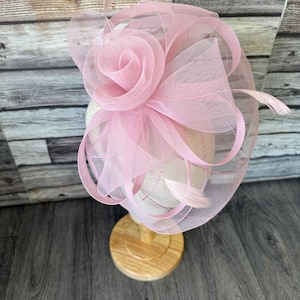 May include: A pale pink fascinator with a large rose and mesh detailing. The headpiece features delicate feathers and is displayed on a wooden stand. The design is suitable for special occasions.