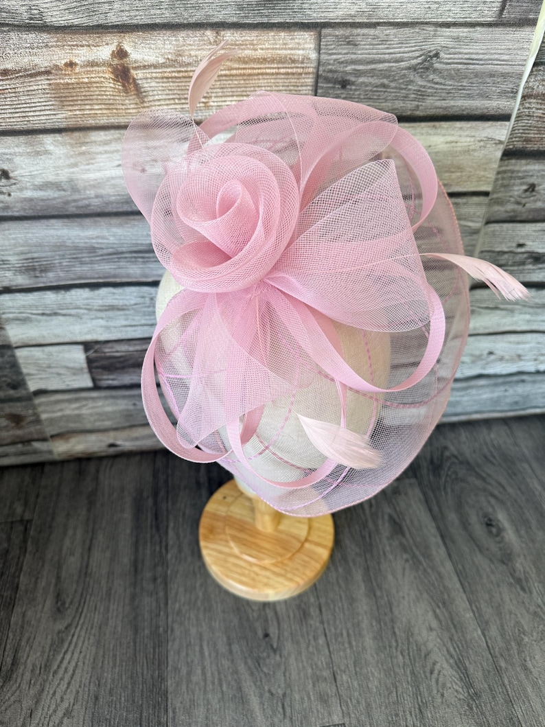 May include: A pale pink fascinator with a large rose and mesh detailing. The headpiece features delicate feathers and is displayed on a wooden stand. The design is suitable for special occasions.
