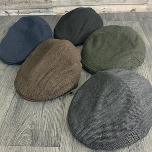 May include: A selection of six flat caps in shades of navy, charcoal, brown and olive green. The caps have a traditional design and are made from textured fabric, with a herringbone pattern on some.