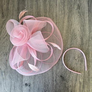 May include: A pale pink fascinator featuring a rose-shaped embellishment, mesh detailing, and feathers. A matching pink headband is also present. Ideal for weddings or formal events.