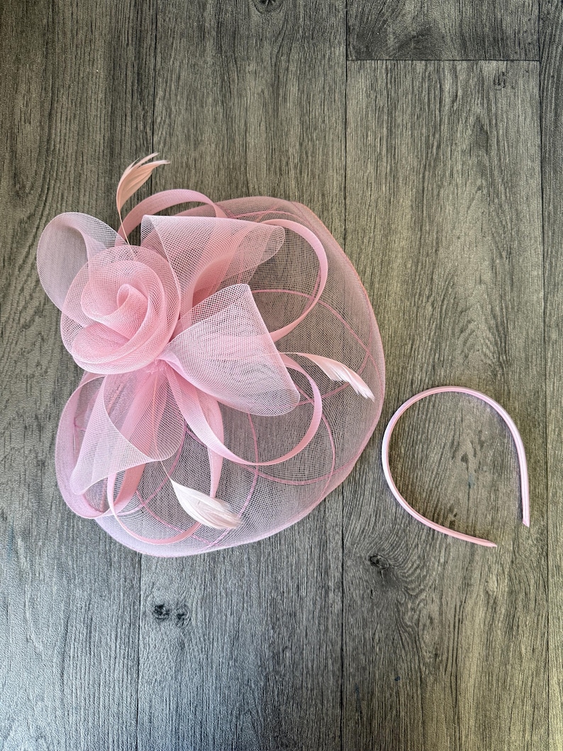 May include: A pale pink fascinator featuring a rose-shaped embellishment, mesh detailing, and feathers. A matching pink headband is also present. Ideal for weddings or formal events.
