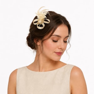 May include: A cream-colored hair clip featuring a flower and feather design. The hair accessory is attached to dark brown hair. The woman is wearing a cream-colored sleeveless dress.