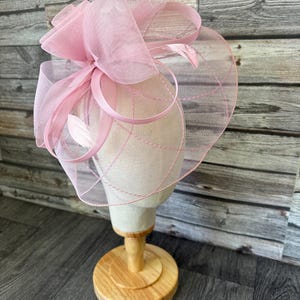 May include: A pale pink fascinator hat with a mesh veil, ribbons, and feathers. The hat is displayed on a mannequin head with a wooden stand. The hat has a circular shape with a large bow on top.