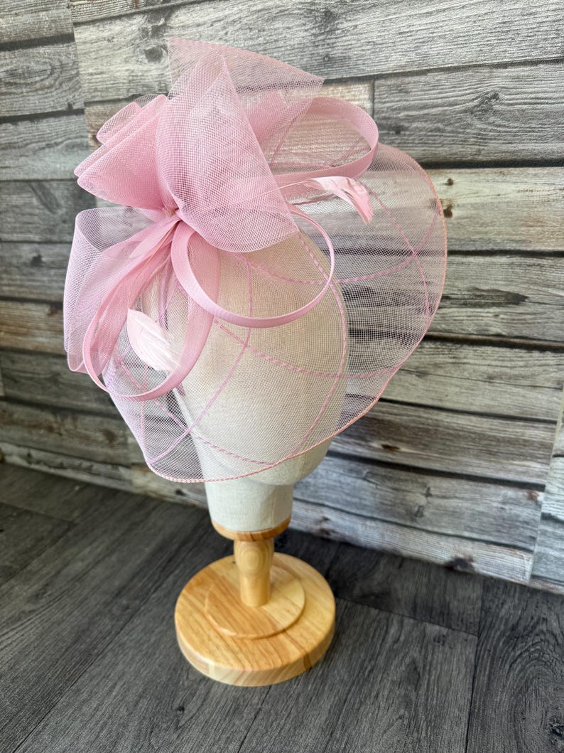 May include: A pale pink fascinator hat with a mesh veil, ribbons, and feathers. The hat is displayed on a mannequin head with a wooden stand. The hat has a circular shape with a large bow on top.