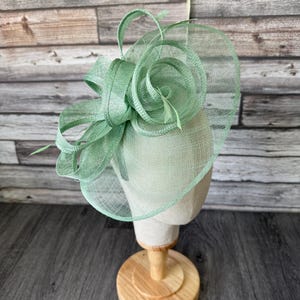 Peut inclure: Un fascinateur vert menthe avec une base circulaire et des ornements floraux. Le chapeau est présenté sur une tête de mannequin, monté sur un support en bois. Le design présente une texture tissée et des lignes élégantes.