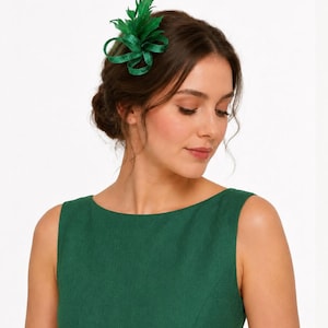 Peut inclure: Accessoire pour cheveux vert avec un nœud et des plumes, épinglé sur le côté des cheveux. La femme porte une robe verte. Le fond est blanc.