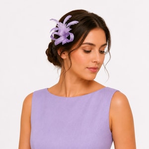 May include: A lavender hair clip featuring feather and ribbon detailing. The hair accessory is positioned on the side of the head. The woman in the image is wearing a lavender dress.