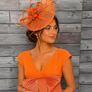 May include: An orange fascinator with a flower and feather accents. The woman is wearing an orange dress and holding an orange clutch. The background is a gray wood-paneled wall. The overall theme is formal wear.