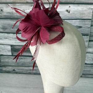 May include: A burgundy fascinator with a flower and feather design. The headpiece features a large, layered flower and several long, slender feathers. A textured ribbon wraps around the base of the flower. The fascinator is displayed on a mannequin head.