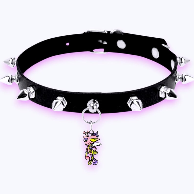 May include: A black collar featuring a series of silver-toned metal spikes along the band and a central silver ring holding a small, colorful cartoon character charm with pink, yellow, and purple details. The collar includes a metal buckle closure.