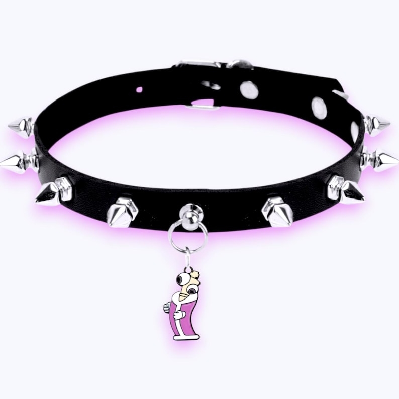May include: A black choker collar featuring silver-toned metal spikes arranged around the band. The collar includes a silver metal buckle closure and a dangling charm depicting a cartoon character with a single eye wearing a pink robe.