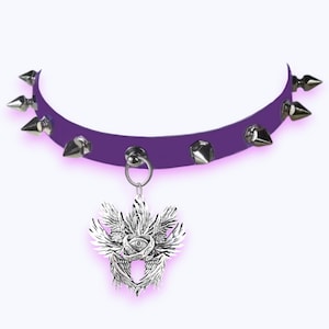 May include: A purple choker necklace with silver spikes and a pendant. The pendant features a detailed design of wings, a rose, and an eye. The necklace has a metal ring for attachment.