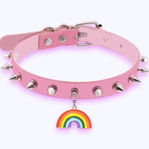May include: A light pink collar featuring a series of silver-toned metal spikes along the band. The collar includes a silver-toned metal buckle and a dangling charm shaped like a colorful rainbow with red, orange, yellow, green, blue, and purple stripes.