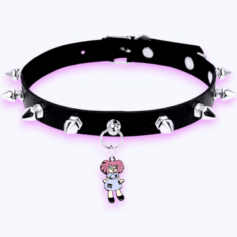 May include: A black choker collar featuring a series of silver-toned metal spikes along the band. A silver-toned metal ring is attached to the front, holding a small, colorful enamel charm depicting a cartoon doll with pink hair and a blue dress.
