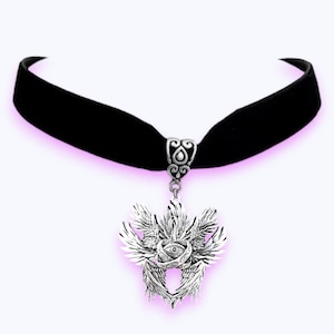 May include: A black velvet choker necklace with a silver pendant. The pendant has a detailed design of wings, an eye, and ornate detailing. The necklace is set against a white background with a subtle pink glow.