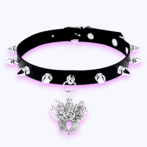 May include: Black leather choker necklace with silver-coloured cone studs and a decorative pendant. The pendant features a detailed design with an eye in the centre, surrounded by wings and ornate details.