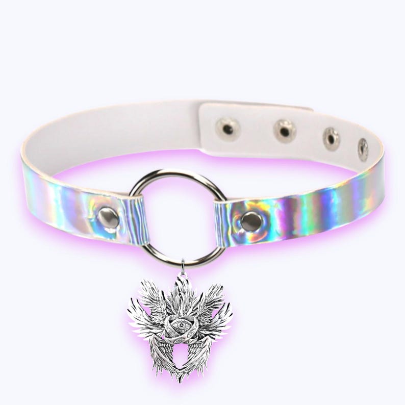 May include: A white choker necklace with a holographic iridescent band. The necklace features a silver metal ring and a pendant with a detailed winged eye design. The choker has adjustable poppers for sizing.
