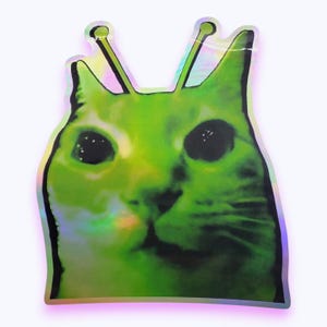 Pode incluir: Um autocolante holográfico com um gato verde brilhante, olhos grandes, escuros e afastados, e duas hastes finas semelhantes a antenas no topo da cabeça. O gato tem um contorno preto definido e um acabamento iridescente e cintilante.