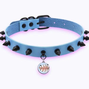 May include: A light blue collar featuring a series of black metal spikes along the band. The collar includes a black buckle closure and a circular charm hanging from the centre, which displays a graphic of a round face with sharp, jagged teeth.