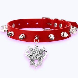 May include: Red leather choker necklace with silver spikes and a pendant. The pendant has a detailed design of wings and an eye. The choker has a buckle closure, a bold accessory.