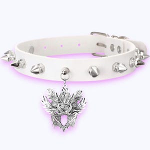 May include: White leather choker necklace with silver spikes and a decorative pendant. The pendant has an eye at the centre, surrounded by wings and patterns. The choker has a buckle fastening.