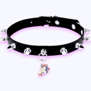 May include: A black choker collar featuring silver-toned metal spikes arranged around the band. The collar includes a central silver-toned ring holding a small, colourful enamel charm depicting a stylised cartoon character with black and red fur.