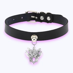 May include: Black leather choker necklace with a silver pendant. The pendant has an eye design at the centre, surrounded by wings and patterns. The choker has a buckle fastening and a metal loop for the pendant.