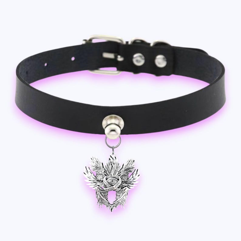 May include: Black leather choker necklace with a silver pendant. The pendant has an eye design at the centre, surrounded by wings and patterns. The choker has a buckle fastening and a metal loop for the pendant.