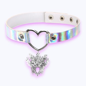 May include: White choker necklace with a silver heart-shaped pendant and a decorative silver charm. The choker has an iridescent band and adjustable snap closures. The charm features a detailed design with an eye and wings.