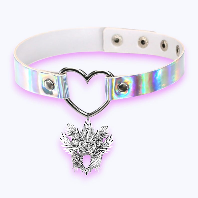 May include: White choker necklace with a silver heart-shaped pendant and a decorative silver charm. The choker has an iridescent band and adjustable snap closures. The charm features a detailed design with an eye and wings.