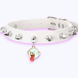 May include: A white choker collar featuring silver-toned metal spikes along the band and a dangling charm shaped like a white ghost with a red tongue. The collar includes a silver-toned metal buckle closure with multiple adjustment holes.